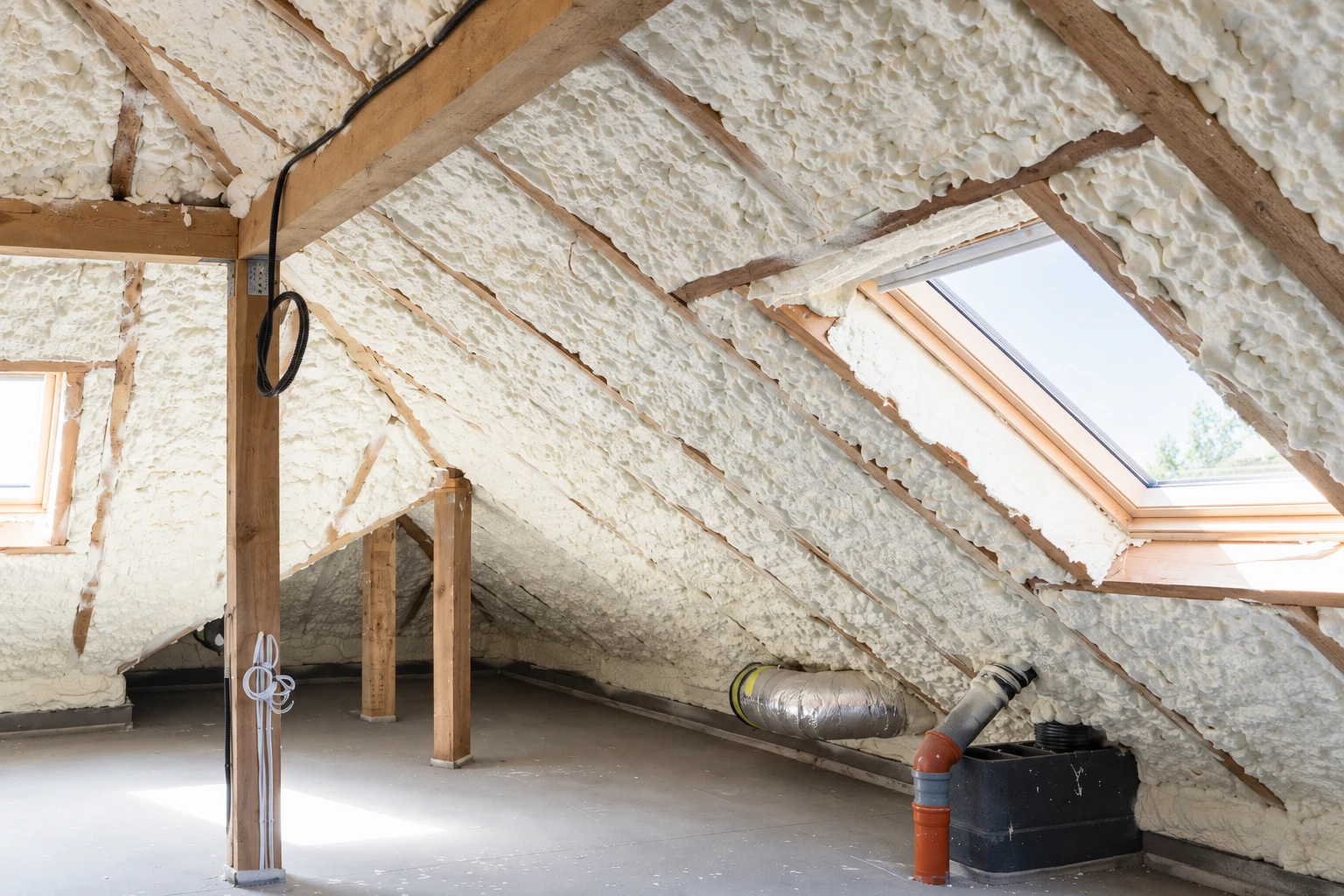 Spray foam insulation covers the sloped roof framing in an unfinished attic with skylights, exposed beams, and ductwork.