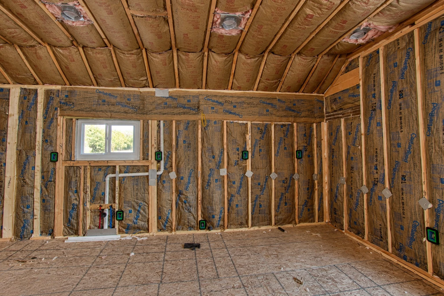 An unfinished attic room has brown fiberglass batts fitted between roof rafters and wall studs around a small window.