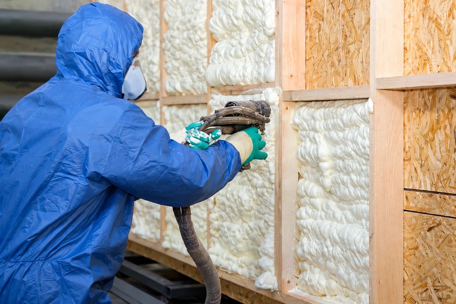 Worker in protective coveralls sprays foam into the bays of a wood-framed wall.