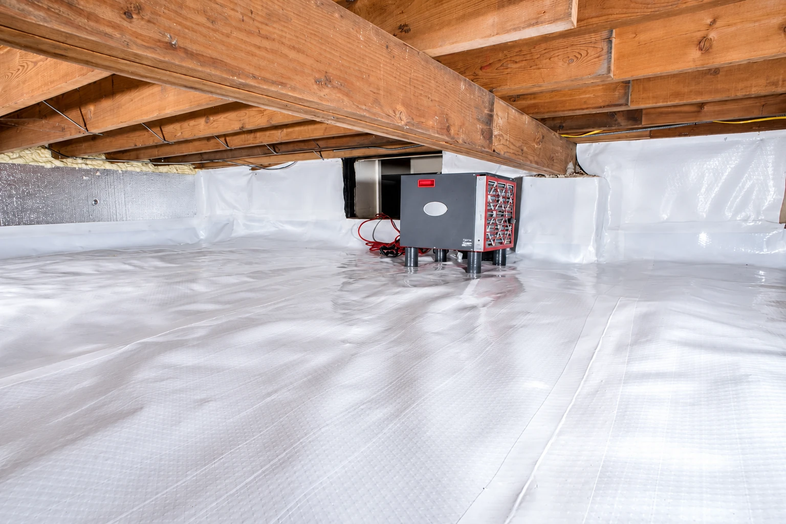 Crawl space with a white vapor barrier covering the floor and walls beneath exposed wooden joists, with a dehumidifier at the back.