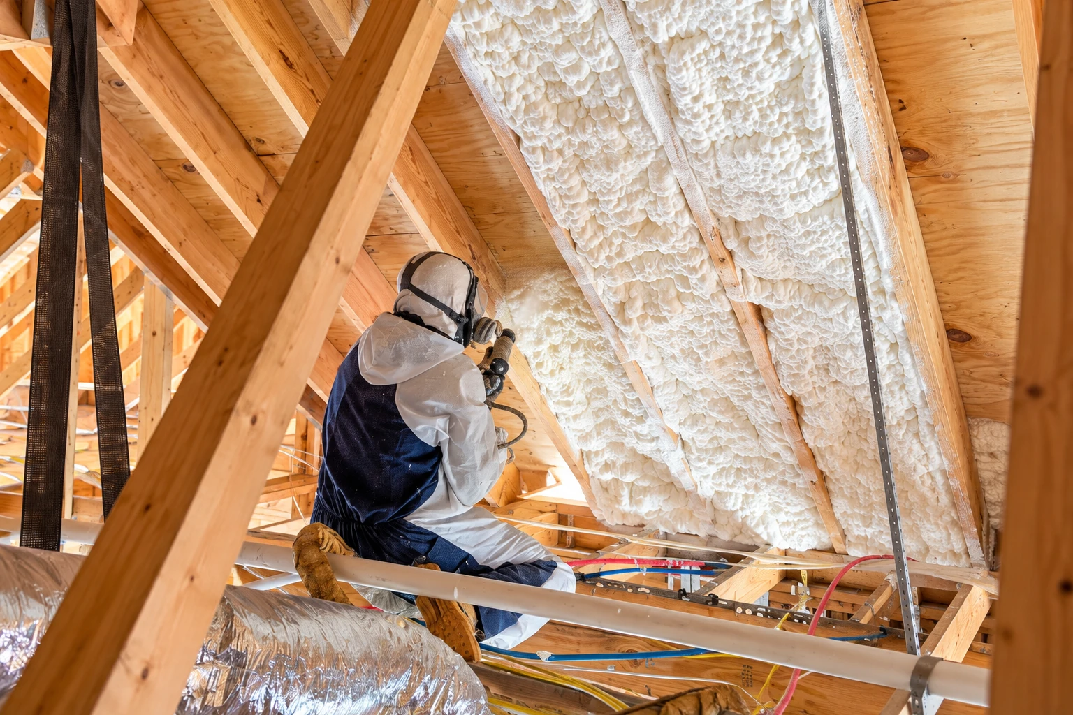 A worker in protective gear sprays white foam insulation between the rafters of an unfinished attic.