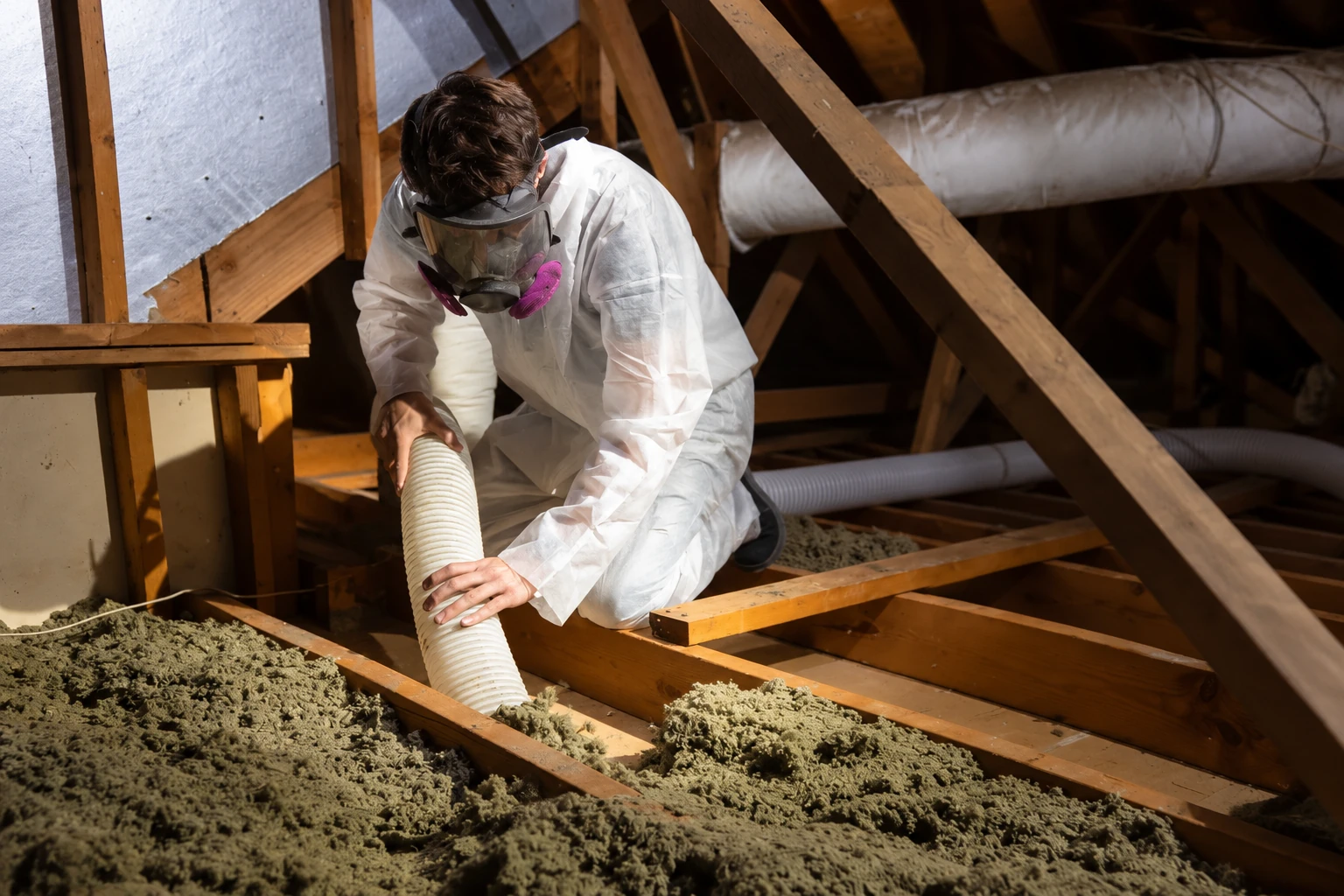 A worker in protective coveralls and a respirator uses a hose to remove old insulation from an attic.