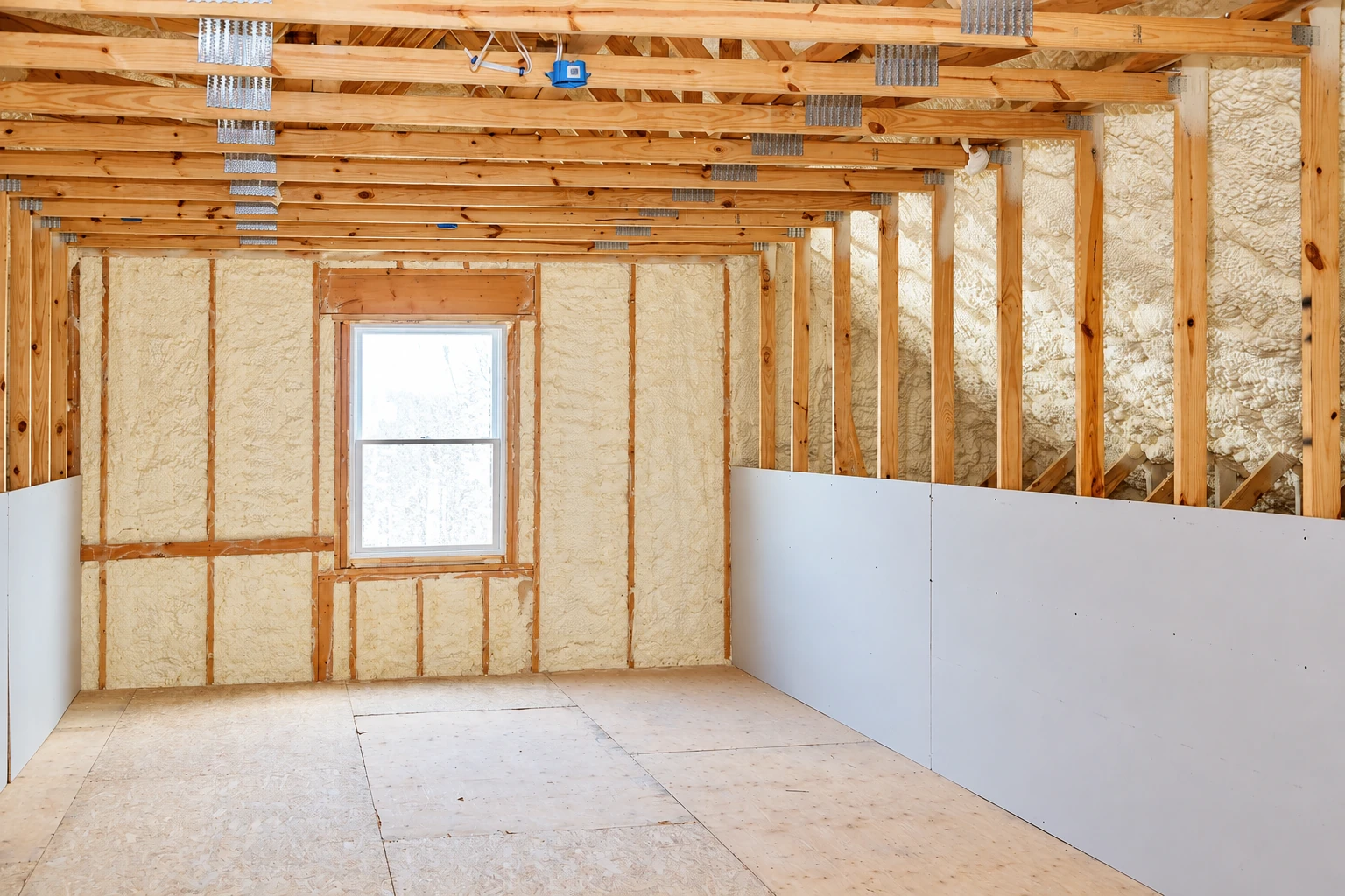 A framed room has pale spray foam filling the wall cavities, with exposed ceiling joists, a window, and partial drywall.